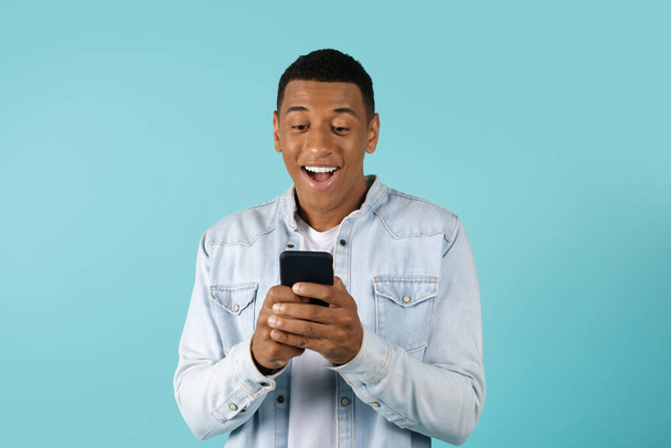 Surprised smiling young african american guy in casual typing on smartphone isolated on blue background. Great ad and offer, app for chat and social networks, good news, technology for communication - Valokuva, kuva