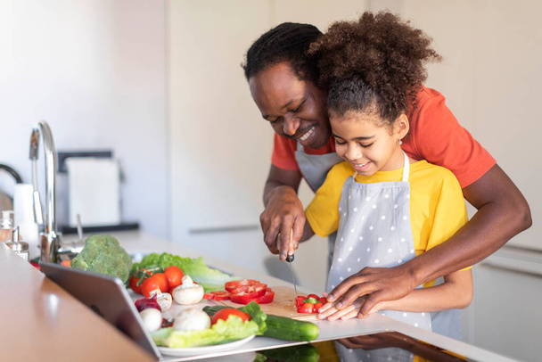 Люблячий чорний батько навчає свою дочку готувати їжу на кухні, щасливий афроамериканський тато і дев'ятнадцять жінок розважаються, готуючи овочевий салат разом вдома, копіювати простір
 - Фото, зображення