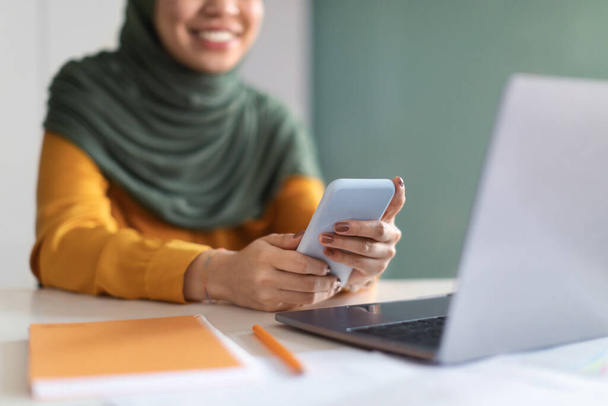 Muslim Woman In Hijab Using Mobile Phone While Sitting At Desk In Office, Unrecognizable Smiling Islamic Lady Shopping Online Or Browsing Modern App For Internet Banking, Closeup With Selective Focus - Фото, зображення