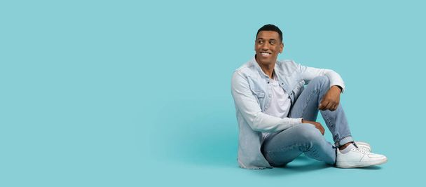 Smiling handsome young african american male in casual sit on floor, look back at free space, enjoy spare time alone isolated on blue background, panorama, studio. Great ad and offer, sale, lifestyle - Foto, Bild