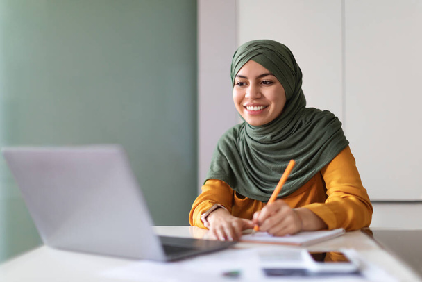 Jeune femme musulmane Regarder Webinaire sur ordinateur portable à la maison et prendre des notes, souriante dame arabe dans Hijab étude en ligne avec ordinateur tout en étant assis au bureau à l'intérieur, profiter de l'enseignement à distance, Espace libre - Photo, image
