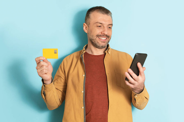 Концепция мобильного шопинга. Счастливый зрелый мужчина, используя смартфон и кредитную карту, делает оплату за синий студийный фон. Банковское приложение и концепция электронной коммерции - Фото, изображение