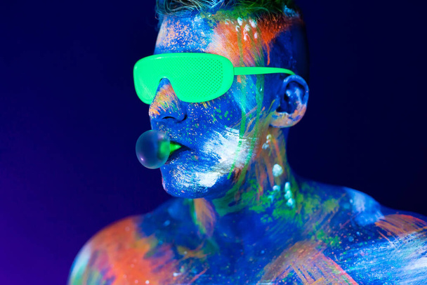 Mladý muž žvýká žvýkačku a nafoukne balón. Zářivka na obličej ve studiu s UV světlem - Fotografie, Obrázek