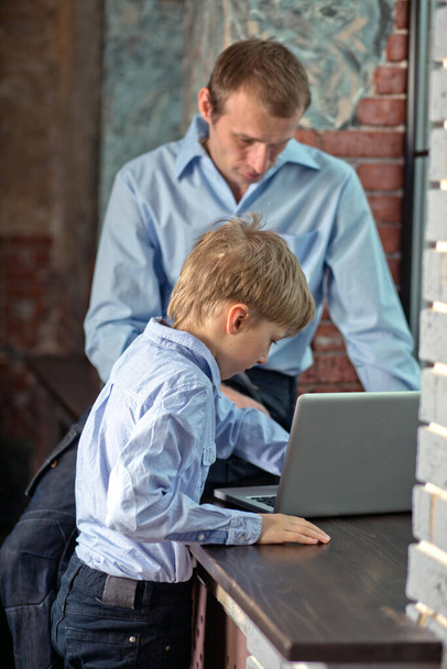 Dad sits on windowsill teaches son to work on computer. Father and son playing on laptop. Father is looking for gift to son in online store in modern loft. Vertical, blurred background. - Photo, Image