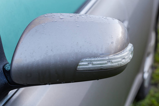 Side view mirror of the parked gray car with integrated turn signal repeater covered with dew, front view on a blurred background of misted side window and door close-up - Photo, Image