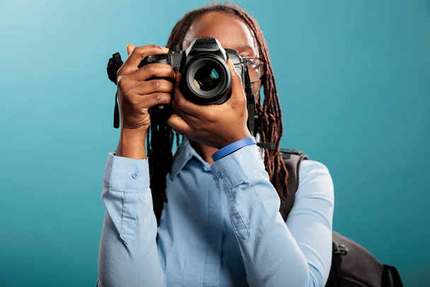 African american young adult photography entusiast with modern photographing device taking photos on blue background. Confident and creative photographer taking pictures while enjoying leisure time. - Φωτογραφία, εικόνα