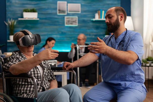Elderly woman uses a virtual reality headset on a wheelchair in a nursing home to explore augmented reality. Male nurse in common room of skilled care facility helping patient to have some fun. - Photo, Image