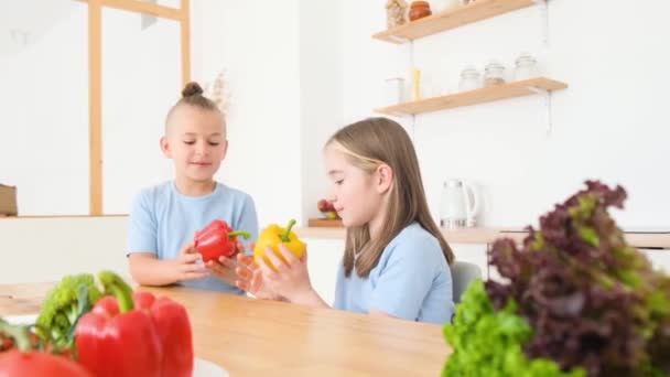 Malý bratr a sestra v modrých tričkách sedí v kuchyni a jedí čerstvou zeleninu. Koncept šťastného dítěte. Zdravé jídlo pro děti - Záběry, video
