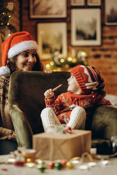 Счастливый ребенок с мамой сидит в кресле в канун Рождества и ребенок ест рождественские конфеты. Декор рождественской комнаты. В ожидании волшебства Рождества. - Фото, изображение