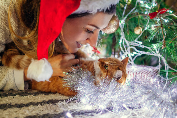 Boże Narodzenie z kotem. Kobieta bawiąca się i przytulająca zwierzaka w kapeluszu Mikołaja przy drzewie w domu. Zwierzęta pokryte błyskotkami i światłami - Zdjęcie, obraz