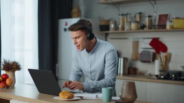Aantrekkelijke vrolijke man dansend op keuken met koptelefoon die muziek luistert op laptop. Leuke jongeman die zingt en geniet van melodie in draadloze headset. Positieve freelancer veel plezier thuis met behulp van technologie. - Video