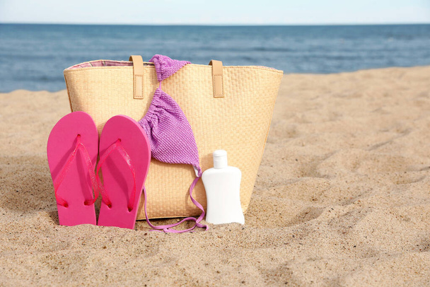 Summer bag with slippers, sunscreen and bikini top on sand near sea, space for text - Fotografie, Obrázek
