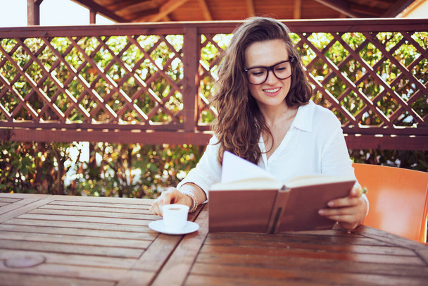 smiling elegant woman in white shirt with eyeglasses sitting at the table drinking coffee and reading book in the terrace of guest house hotel. - Photo, image