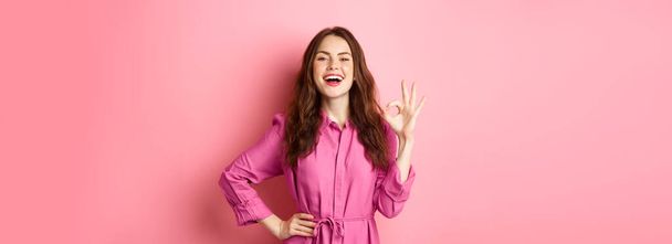 Νεαρή ελκυστική γυναίκα λέει ναι, δείχνει εντάξει σημάδι στην έγκριση, χαμογελώντας σίγουροι και ικανοποιημένοι, στέκεται πάνω από ροζ φόντο. - Φωτογραφία, εικόνα