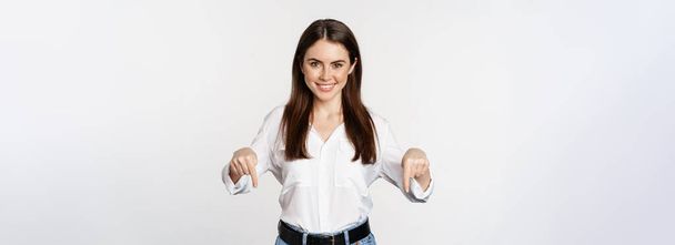 Confident smiling woman pointing fingers down, showing logo, banner below, recommending click on link, standing over white background. - Foto, Imagem