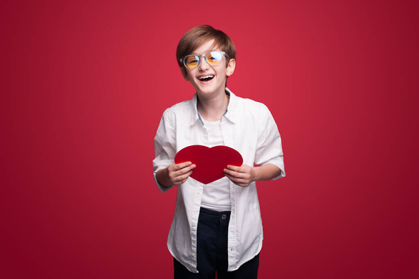 Усміхнений хлопчик у окулярах тримає серце валентинки, вирізане з червоного паперу на червоному тлі. Концепція дня матері. Безтурботне дитинство. Свято фону
. - Фото, зображення