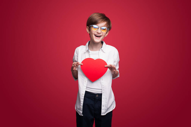 Portret van een blank jongetje met een rood papieren hart op de borst over een rode achtergrond. Positief persoon. Geluksconcept. Geïsoleerd portret. Moederdag concept - Foto, afbeelding