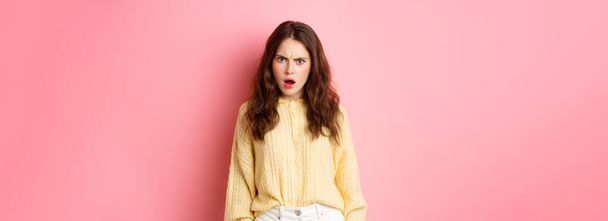Shocked and confused brunette woman gasping, frowning and looking offended at camera, feeling insulted or displeased, standing over pink background. - Photo, Image