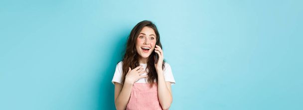 Mooi meisje praten op smartphone en ontvang verrassend nieuws, kijken opgewonden en verbaasd, praten over de telefoon, staande gelukkig tegen een blauwe achtergrond. - Foto, afbeelding