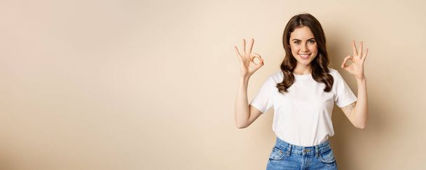 Beautiful female model smiling pleased, showing okay, zero sign, complimenting, praising you, standing over beige background. - Foto, afbeelding