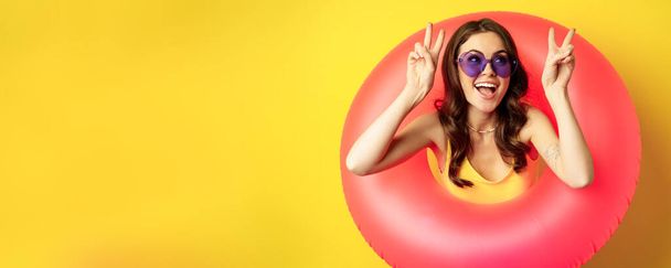 Portrait of happy stylish girl in sunglasses, inside beach swimming ring, showing peace sign, laughing and smiling, standing over yellow background. - Photo, Image