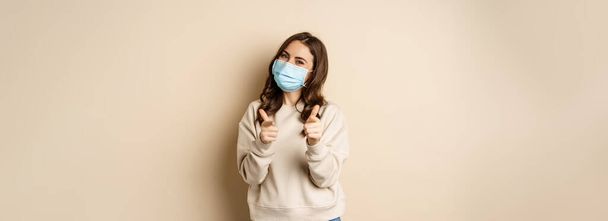 Hey you congrats. Smiling beautiful woman in medical face mask, pointing fingers at camera, standing over beige studio background. - Fotografie, Obrázek