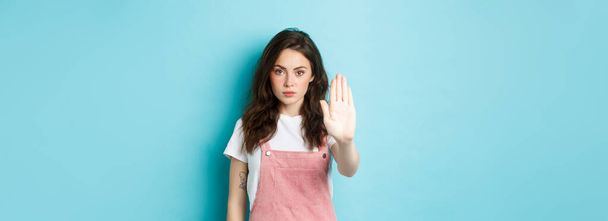 Νεαρή μελαχρινή γυναίκα με σγουρό χτένισμα, σηκώνεται χέρι με χέρι με χειρονομία, λέει στοπ ή όχι, αρνείται κακή προσφορά, απορρίπτει κάτι, στέκεται πάνω σε μπλε φόντο. - Φωτογραφία, εικόνα