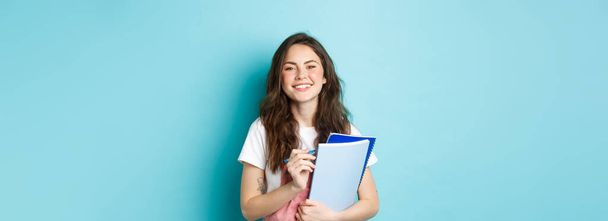 Ευτυχισμένη νεαρή φοιτήτρια που κρατάει σημειωματάρια από τα μαθήματα και χαμογελά στην κάμερα, στέκεται με ανοιξιάτικα ρούχα σε μπλε φόντο. - Φωτογραφία, εικόνα