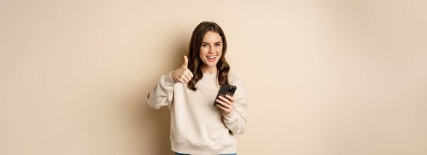 Young woman smiling, showing thumbs up while using mobile phone, smartphone app, standing over beige background. - Foto, imagen
