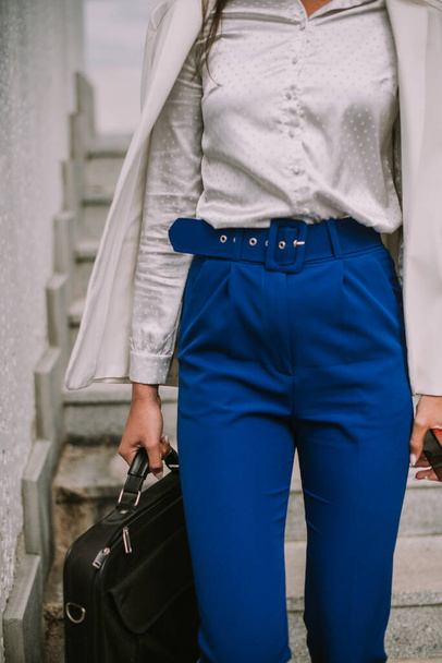 Közelkép egy üzletasszonyról, fehér öltönyben és kék nadrágban, amint lefelé sétál a lépcsőn a bőröndjével. - Fotó, kép