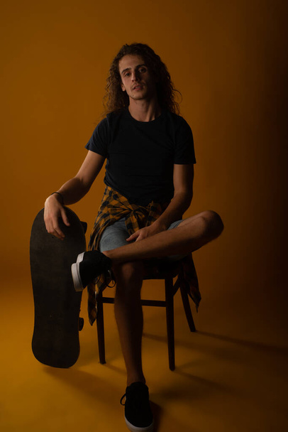 Νεαρός όμορφος άντρας κάθεται σε μια καρέκλα και ξεκουράζει το χέρι του στο σκέιτμπορντ στο στούντιο - Φωτογραφία, εικόνα