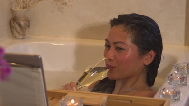 CLOSE UP: Dame im schäumenden Bad trinkt ein Glas Champagner bei einem romantischen Online-Date. Schöne philippinische Frau im entspannenden Schaumbad, lächelnd und flirtend mit Ehemann per Videoanruf auf digitalem Tablet - Filmmaterial, Video