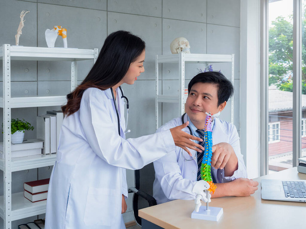 Két fehér egyenruhás ázsiai orvos beszél és dolgozik együtt az orvosi rendelőben. Az ortopéd felnőtt férfi orvos nyújt ismereteket és tanácsokat a női orvos a gerinc az asztalon a kórházban. - Fotó, kép