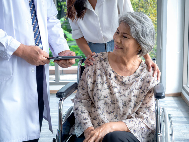 Porträt einer lächelnden älteren Patientin im Rollstuhl mit Assistentin, die sie zum Arzt führt, um ihre Krankenakte zu besprechen. Konzept zur Vorsorgeuntersuchung für Senioren. - Foto, Bild