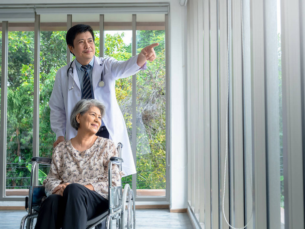 Uśmiechnięta Azjatka seniorka siedzi na wózku inwalidzkim, podczas gdy lekarz w białym płaszczu stoi w gabinecie medycznym w szpitalu, w stylu pionowym. Opieka, opieka zdrowotna i koncepcja medyczna. - Zdjęcie, obraz