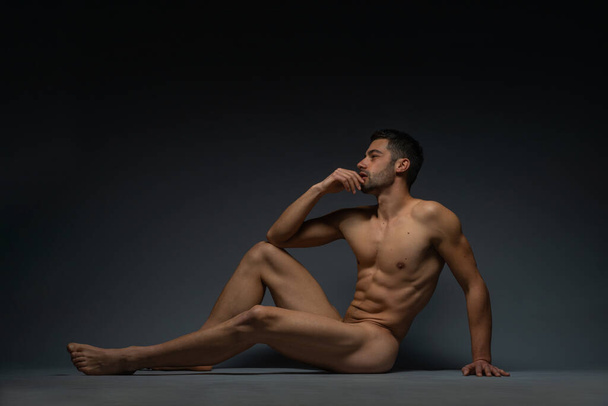 Γυμνή φωτογραφία ανδρικού μοντέλου με σαγηνευτική φιγούρα να κάθεται απομονωμένη στο πάτωμα ενός στούντιο - Φωτογραφία, εικόνα