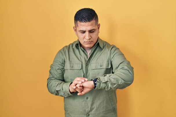 Spanischer junger Mann, der über gelbem Hintergrund steht und die Uhrzeit an der Armbanduhr überprüft, entspannt und selbstbewusst  - Foto, Bild