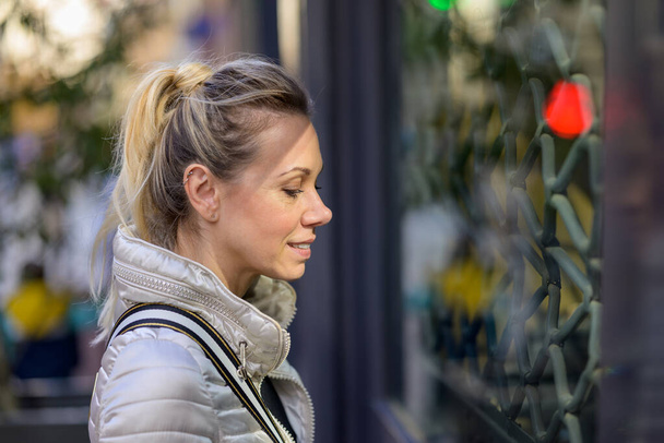 Zijaanzicht van een vrolijke glimlachende vrouw van middelbare leeftijd met blond haar en een paardenstaart die geïnteresseerd is door het raam van een winkel in een winkelstraat - Foto, afbeelding