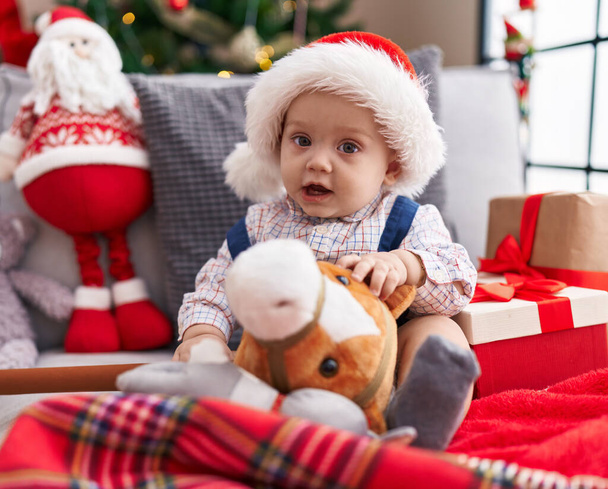 Adorable caucasian baby holding horse toy sitting on sofa by christmas tree at home - Foto, imagen