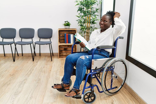Νεαρή μαύρη γυναίκα κάθεται σε αναπηρική καρέκλα στην αίθουσα αναμονής χαμογελώντας αυτοπεποίθηση αγγίζοντας τα μαλλιά με το χέρι προς τα πάνω χειρονομία, θέτοντας ελκυστική και μοντέρνα  - Φωτογραφία, εικόνα