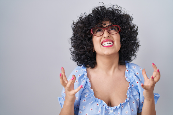Νεαρή μελαχρινή γυναίκα με σγουρά μαλλιά φορώντας γυαλιά σε απομονωμένο φόντο τρελή και τρελή φωνάζοντας και φωνάζοντας με επιθετική έκφραση και χέρια υψωμένα. έννοια απογοήτευσης.  - Φωτογραφία, εικόνα