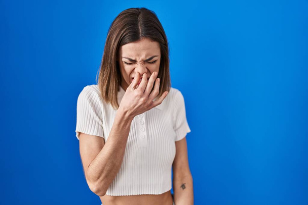 Femme hispanique debout sur fond bleu sentant quelque chose de puant et dégoûtant, odeur intolérable, retenant souffle avec les doigts sur le nez. mauvaise odeur  - Photo, image
