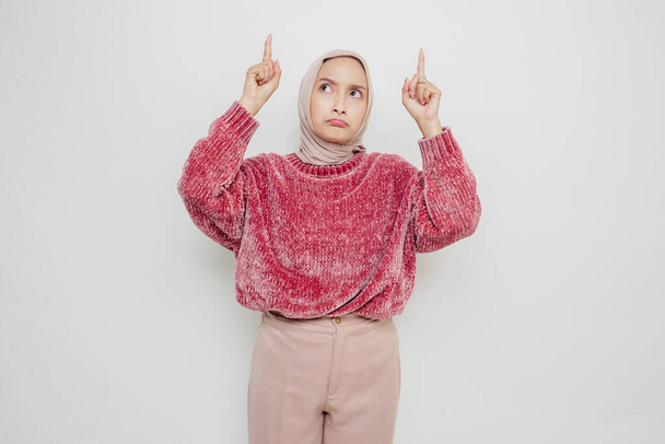 Shocked Asian Muslim woman wearing pink sweater and hijab pointing at the copy space on top of her, isolated by white background - Foto, immagini