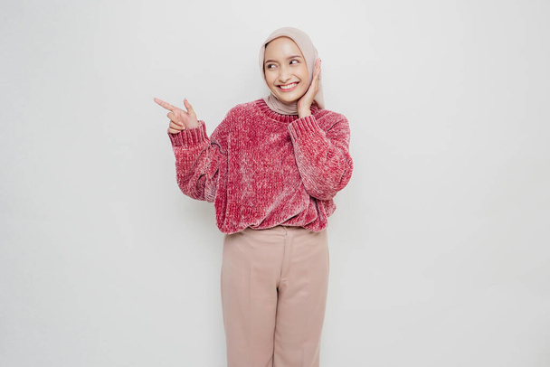 Excited Asian Muslim woman wearing pink sweater and hijab pointing at the copy space beside her, isolated by white background - Φωτογραφία, εικόνα