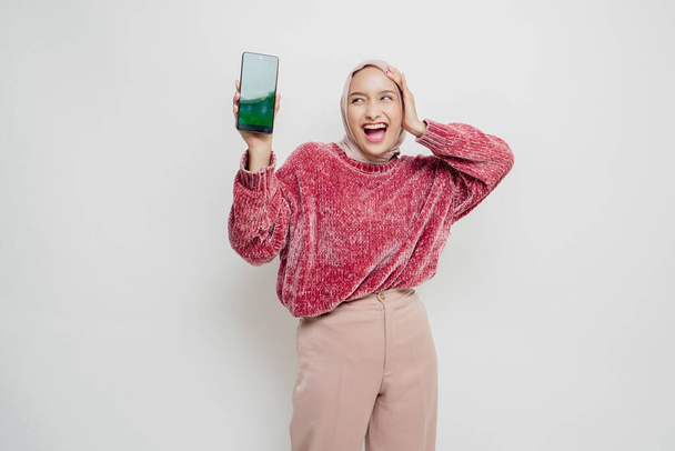 Izgalmas ázsiai muszlim nő rózsaszín pulóvert és hidzsábot visel, miközben a mellette lévő fénymásolótérre mutat, miközben a telefonját tartja, fehér háttérrel elszigetelve. - Fotó, kép