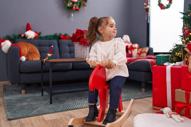 Adorable hispanic girl playing with reindeer rocking by christmas tree at home - Foto, imagen