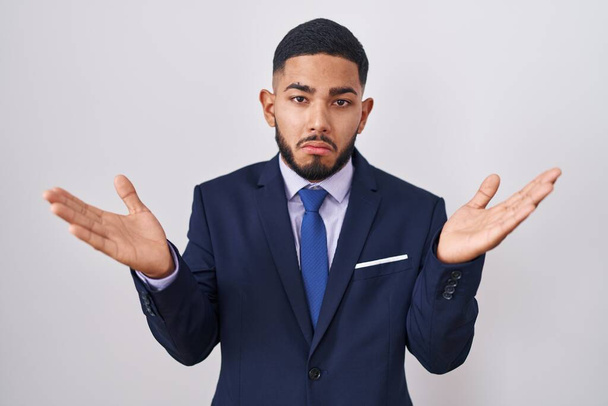 Young hispanic man wearing business suit and tie clueless and confused expression with arms and hands raised. doubt concept.  - Photo, Image