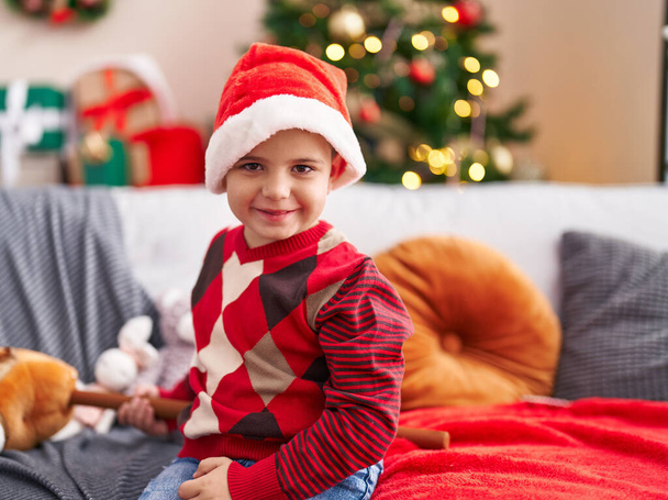 Adorable niño hispano sonriendo confiado sentado en el sofá por el árbol de Navidad en casa - Foto, imagen