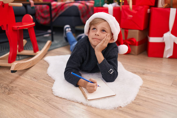 Adorable toddler writing on notebook lying by christmas tree at home - 写真・画像