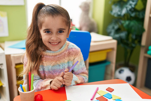 Αξιολάτρευτο ισπανόφωνο κορίτσι φοιτητής χαμογελά αυτοπεποίθηση κρατώντας μολύβι χρώμα στο νηπιαγωγείο - Φωτογραφία, εικόνα